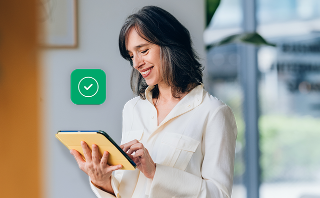 A person in a light-colored blouse uses a tablet indoors while a green checkmark icon appears beside them, suggesting a successful action or approval.