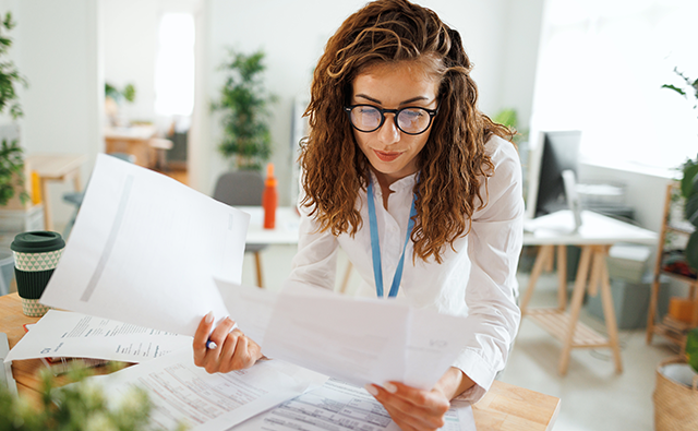 A person wearing glasses reviews printed documents spread across a desk in a bright modern office with plants and workstations in the background.