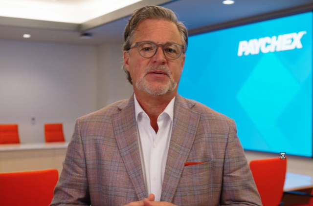 A person wearing glasses, a white shirt, and a plaid blazer stands in a modern conference room, with a large screen displaying the Paychex logo and bright orange chairs in the background.