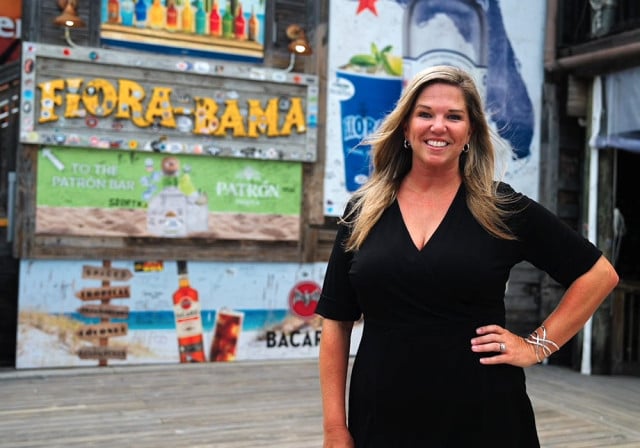 A person in a black dress stands on a wooden deck in front of colorful beverage advertisements and a rustic sign reading “Flora-Bama.”