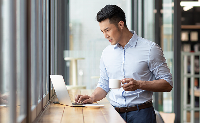 A person standing by a window works on a laptop while holding a coffee cup in a bright modern office.