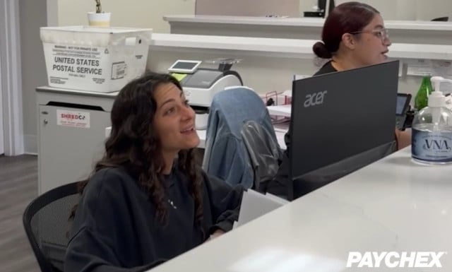 Two office employees sit behind a reception desk with computer monitors, office equipment, a USPS mail bin, and a hand sanitizer bottle visible in a modern workplace.