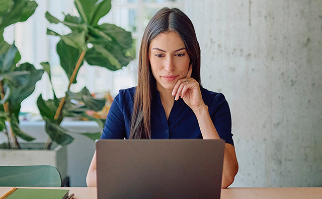Person seated at a desk in a modern office, thoughtfully looking at a laptop computer with a large green plant in the background.