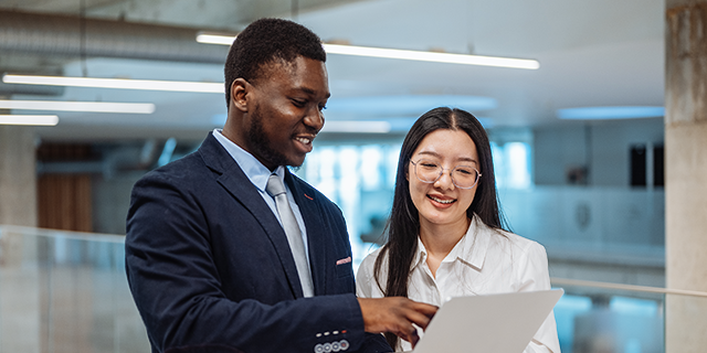 Two people in business attire review content on a laptop together in a modern office environment.
