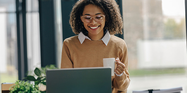 Woman with glasses holding white mug, positioned in front of window, looks at her computer